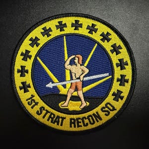 May include: Embroidered patch with a circular design. The patch features a central blue field with a figure holding a spear, surrounded by yellow rays. The text "1st STRAT RECON SQ" is in yellow, with a yellow border.