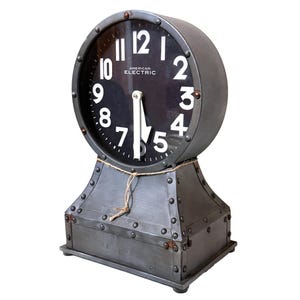 May include: A dark gray metal clock with a round face, white numerals, and the text AMERICAN ELECTRIC. The clock features a riveted base and a circular housing, with a piece of twine tied around the neck connecting the base to the clock face.