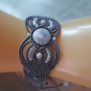 May include: A gray macrame bracelet featuring a large, round, white stone with gray veining at the center, flanked by smaller white stones and white beads. The bracelet is handmade with intricate knotting and a woven design, showcasing a bohemian style.