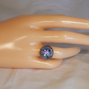 Puede incluir: Un anillo redondo plateado con una cúpula de resina transparente teñida de azul. Dentro de la cúpula hay piezas de purpurina iridiscente con forma de copo de nieve. El anillo se muestra en una mano clara.
