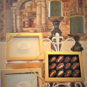 Puede incluir: Una pila de cajas doradas con una cinta dorada atada alrededor. Las cajas están llenas de dátiles y nueces. Las cajas están sobre una superficie blanca frente a una pintura de una ciudad.