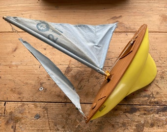 May include: A vintage toy sailboat with a yellow hull and wooden deck. The light blue sails have the number "21" and an anchor symbol. The word "GULL" is printed on the side of the hull. The boat is set against a wooden background.