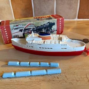 Può includere: Un'antica nave giocattolo con scafo rosso, sovrastruttura bianca e fumaiolo blu, arancione e bianco. Il giocattolo è accompagnato da una scatola e due file di piccoli vagoni blu. La scatola presenta illustrazioni di una nave e di un treno.