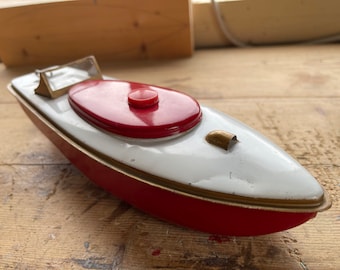 May include: A vintage toy boat with a red and white color scheme. The boat features a red oval-shaped top, a small clear windshield, and gold-colored trim. The boat is made of metal and sits on a wooden surface.