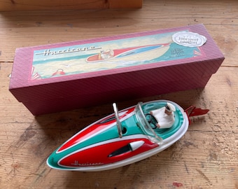 May include: Vintage toy boat with a red, white, and teal color scheme, featuring a driver figure. The boat is next to a maroon box with a boat illustration and the word "Hurricane".