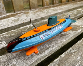 May include: A vintage toy submarine in blue, orange, and green, with the words "GYRO MARINER" printed on the side. The toy has a silver antenna and orange fins. The submarine is resting on a wooden surface.