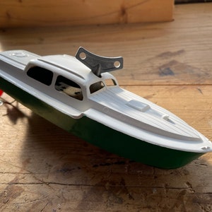 Puede incluir: Un barco de juguete vintage con cabina blanca y casco verde. El barco tiene una llave de cuerda y una pequeña hélice roja. La cubierta es blanca con detalles moldeados. El juguete está sobre un fondo de madera.