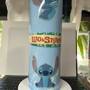K&ouml;nnte beinhalten: Ein hellblauer Becher mit der Figur Stitch aus Lilo & Stitch. Der Becher hat zwei Bilder von Stitch, den Text "Lilo & Stitch" und ein Blumenmuster. Der Becher ist zylindrisch und scheint aus Metall zu sein.