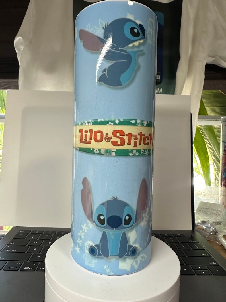 K&ouml;nnte beinhalten: Ein hellblauer Becher mit der Figur Stitch aus Lilo & Stitch. Der Becher hat zwei Bilder von Stitch, den Text "Lilo & Stitch" und ein Blumenmuster. Der Becher ist zylindrisch und scheint aus Metall zu sein.