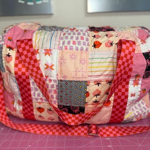 Peut inclure: Un sac polochon matelassé avec un motif patchwork. Le sac présente une variété de carrés de tissu roses, blancs et noirs avec des motifs de fraises et d'étoiles. Les sangles sont à carreaux rouges et roses.