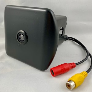 May include: A black rear view camera with a round lens and a black housing. The camera is connected to a red and yellow RCA cable.