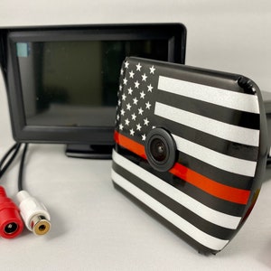 May include: A black and white American flag with a red stripe across the bottom, featuring a small black camera lens in the center. The camera is connected to a black monitor with a screen.