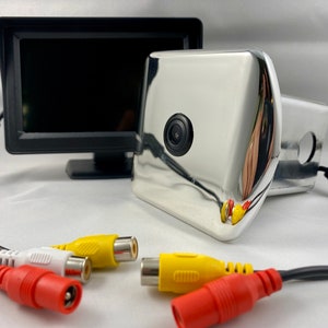 Puede incluir: Un sistema de cámara de marcha atrás cromado con un pequeño monitor negro y cables RCA de colores. La cámara tiene una lente y está montada en una carcasa curva y rectangular. El monitor es negro y rectangular.
