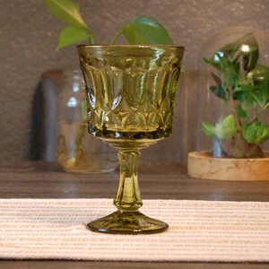Puede incluir: Copa de vino verde oliva de estilo vintage con un diseño estampado. El vaso tiene un tallo y una base ancha. La copa está sobre un mantel individual a rayas beige y blanco. Se ven plantas en el fondo.