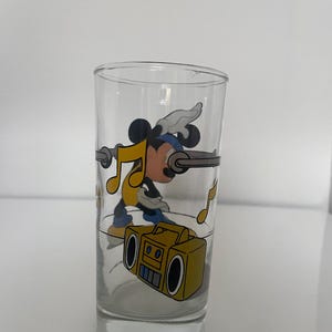 May include: A clear glass tumbler featuring a colorful graphic of a cartoon mouse wearing a blue headband and yellow shirt, dancing near a yellow boombox. The design includes floating yellow musical notes and a horizontal gray bar across the glass.