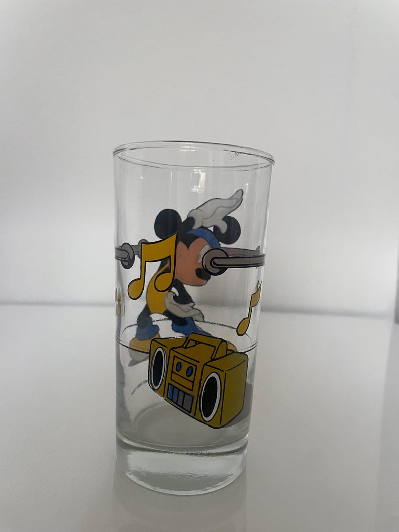 May include: A clear glass tumbler featuring a colorful graphic of a cartoon mouse wearing a blue headband and yellow shirt, dancing near a yellow boombox. The design includes floating yellow musical notes and a horizontal gray bar across the glass.