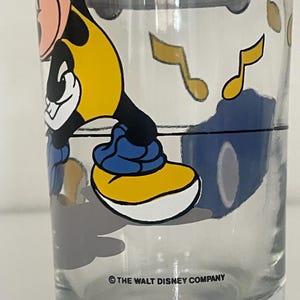 May include: A clear glass tumbler featuring a graphic of Mickey Mouse wearing a yellow shirt and blue shoes. The design includes floating yellow musical notes and a black horizontal line. The text &copy; THE WALT DISNEY COMPANY is printed at the base.