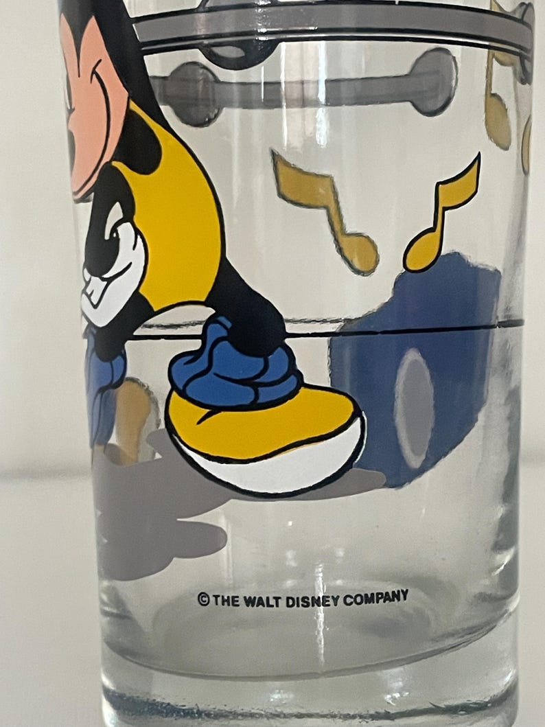 May include: A clear glass tumbler featuring a graphic of Mickey Mouse wearing a yellow shirt and blue shoes. The design includes floating yellow musical notes and a black horizontal line. The text &copy; THE WALT DISNEY COMPANY is printed at the base.