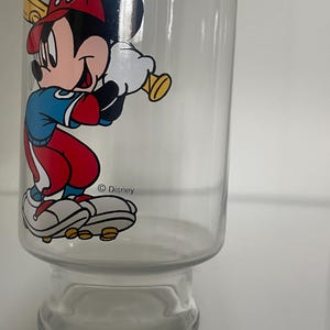 May include: A clear glass tumbler featuring a colorful graphic of Mickey Mouse dressed in a baseball uniform with a red cap, blue shirt, and red pants, holding a yellow baseball bat. The text Disney is printed in black near the character's feet.