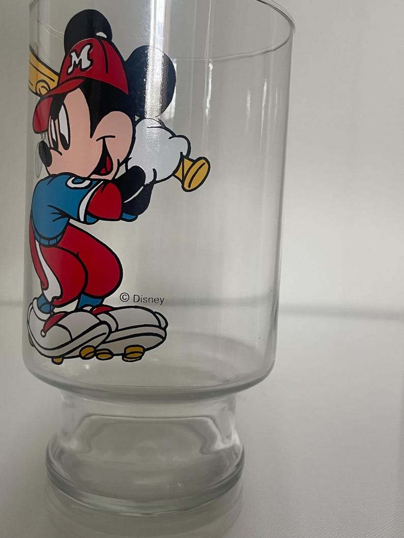 May include: A clear glass tumbler featuring a colorful graphic of Mickey Mouse dressed in a baseball uniform with a red cap, blue shirt, and red pants, holding a yellow baseball bat. The text Disney is printed in black near the character's feet.