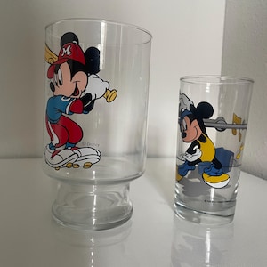 May include: Two clear glass tumblers featuring Mickey Mouse graphics. The larger glass shows Mickey in a red and blue baseball uniform holding a bat. The smaller glass depicts Mickey in a yellow and black outfit with musical notes nearby. Both glasses feature the Disney logo.