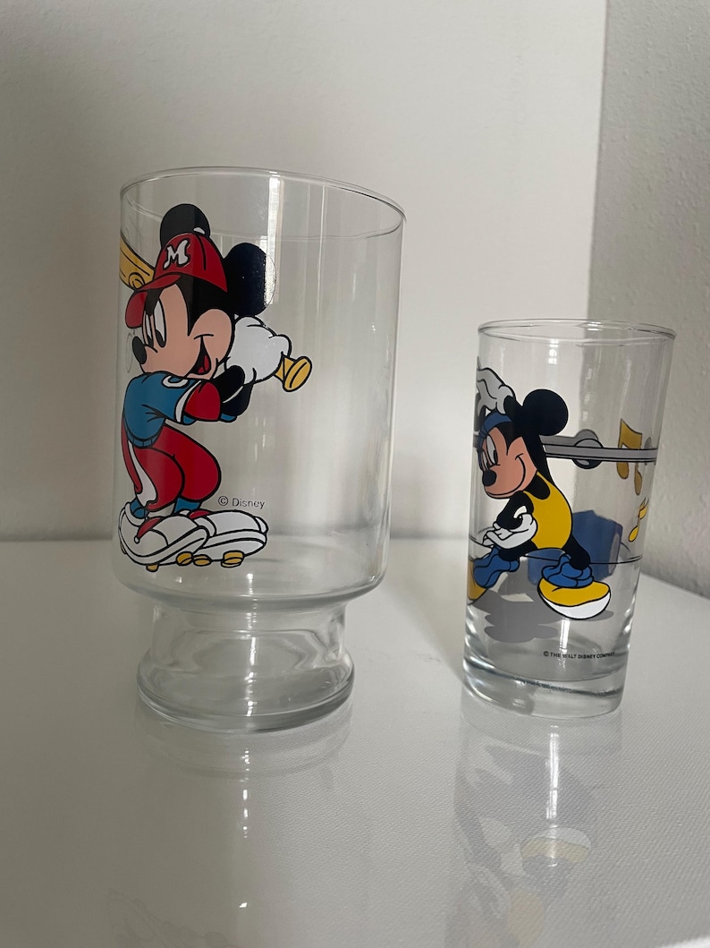 May include: Two clear glass tumblers featuring Mickey Mouse graphics. The larger glass shows Mickey in a red and blue baseball uniform holding a bat. The smaller glass depicts Mickey in a yellow and black outfit with musical notes nearby. Both glasses feature the Disney logo.