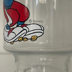 May include: A clear glass tumbler featuring a colorful graphic of a cartoon character wearing a blue shirt, red pants, and white sneakers with yellow soles. The graphic includes the text Disney printed in black ink near the character's feet.