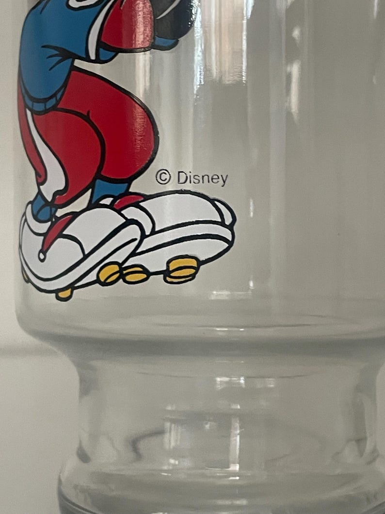 May include: A clear glass tumbler featuring a colorful graphic of a cartoon character wearing a blue shirt, red pants, and white sneakers with yellow soles. The graphic includes the text Disney printed in black ink near the character's feet.