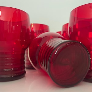 Könnte beinhalten: Eine Sammlung leuchtend roter Trinkgläser. Die Gläser haben eine abgerundete Form mit einem dicken Boden und horizontalen Rillen. Sie bestehen aus einem glänzenden, transparenten Material, das das Licht reflektiert. Ein Blickfang für jede Tischdekoration.