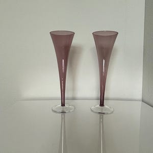 May include: Two tall, slender champagne flutes featuring a muted mauve-colored glass bowl that tapers down to a clear, circular glass base. The glasses have a smooth, minimalist silhouette with a long, thin stem integrated into the bowl design.