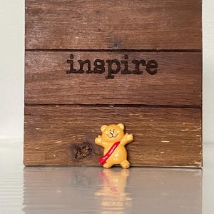 Könnte beinhalten: Ein quadratisches Holzschild aus horizontalen Brettern mit dem eingravierten Wort inspire in der Mitte. Eine kleine gelbe Bärenfigur mit einer roten Schärpe, auf der 100% HUGGABLE steht, ist unten in der Mitte des Holzschildes platziert.