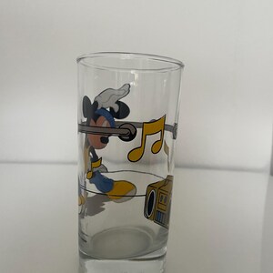 May include: A clear glass tumbler featuring a graphic of Mickey Mouse wearing a blue hat and yellow shoes, listening to a yellow boombox. The design includes floating yellow musical notes and a horizontal gray line encircling the glass.