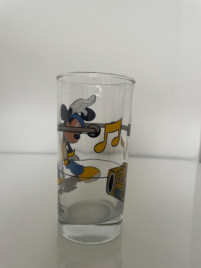 May include: A clear glass tumbler featuring a graphic of Mickey Mouse wearing a blue hat and yellow shoes, listening to a yellow boombox. The design includes floating yellow musical notes and a horizontal gray line encircling the glass.