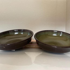 May include: Two round ceramic bowls with a dark brown exterior and a glossy, mottled olive green interior. The bowls feature a smooth, curved rim and a deep basin, showcasing a distinct colour contrast between the dark outer walls and the lighter green centre.