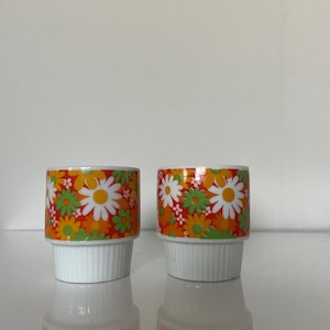 Puede incluir: Dos hueveras cilíndricas con un vibrante estampado floral de margaritas blancas, naranjas y verdes sobre un fondo rojo. La base de cada huevera es blanca con una textura acanalada vertical, lo que crea un contraste con la parte superior.