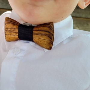 Puede incluir: Una pajarita de madera con una cinta negra. La pajarita est&aacute; hecha de madera marr&oacute;n claro con vetas marr&oacute;n oscuro. La pajarita est&aacute; atada alrededor del cuello de una camisa blanca.