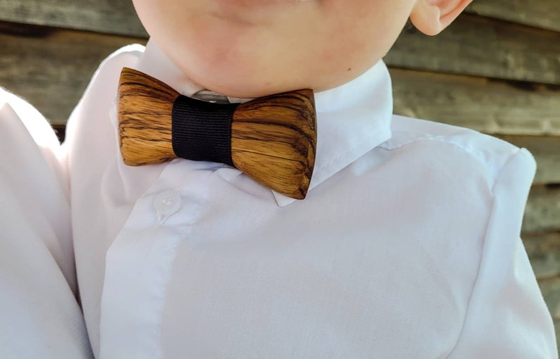 Puede incluir: Una pajarita de madera con una cinta negra. La pajarita est&aacute; hecha de madera marr&oacute;n claro con vetas marr&oacute;n oscuro. La pajarita est&aacute; atada alrededor del cuello de una camisa blanca.