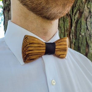 Puede incluir: Una pajarita de madera con una cinta negra. La pajarita est&aacute; hecha de madera oscura con un patr&oacute;n de veta natural.