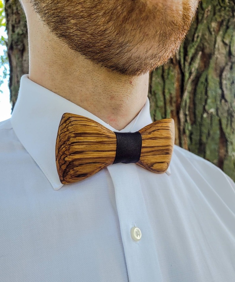 Puede incluir: Una pajarita de madera con una cinta negra. La pajarita est&aacute; hecha de madera oscura con un patr&oacute;n de veta natural.