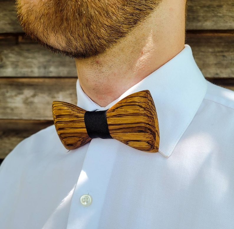 Puede incluir: Una pajarita de madera con una banda negra. La pajarita est&aacute; hecha de madera marr&oacute;n oscura con un patr&oacute;n de veta natural.