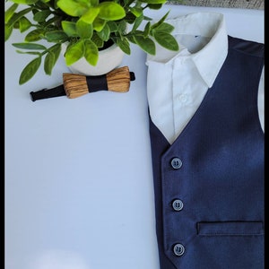 Puede incluir: Un chaleco azul marino con cuatro botones negros y un bolsillo. El chaleco se combina con una camisa blanca de cuello y una pajarita de madera.