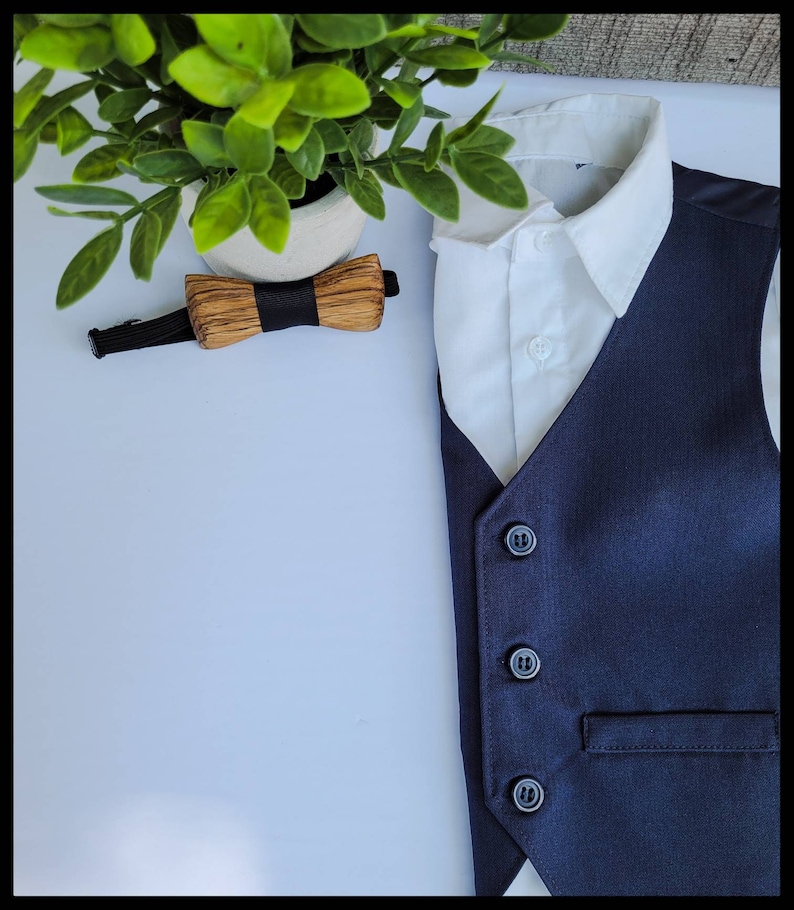 Puede incluir: Un chaleco azul marino con cuatro botones negros y un bolsillo. El chaleco se combina con una camisa blanca de cuello y una pajarita de madera.