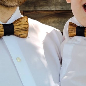 Puede incluir: Dos personas con camisas blancas y pajaritas de madera. Las pajaritas son marrones con ribetes negros.