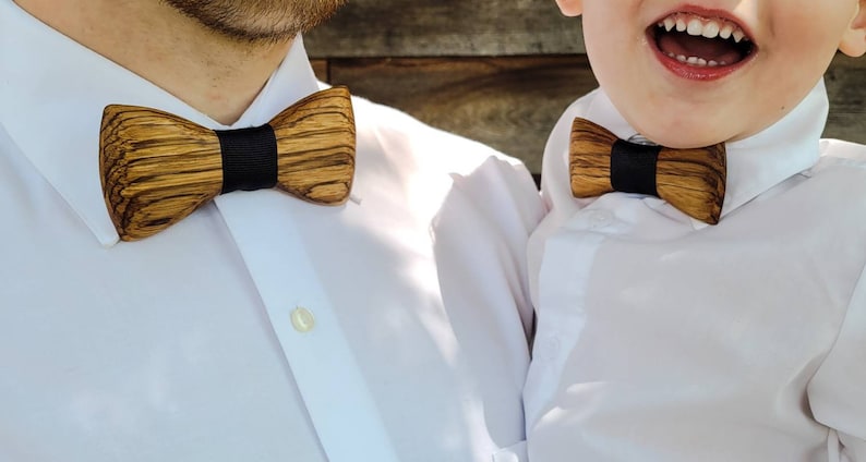 Puede incluir: Dos personas con camisas blancas y pajaritas de madera. Las pajaritas son marrones con ribetes negros.