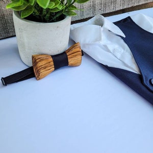 Puede incluir: Una pajarita de madera con una banda el&aacute;stica negra. La pajarita est&aacute; hecha de madera marr&oacute;n claro y tiene una veta de madera natural. La pajarita est&aacute; sobre una superficie blanca.