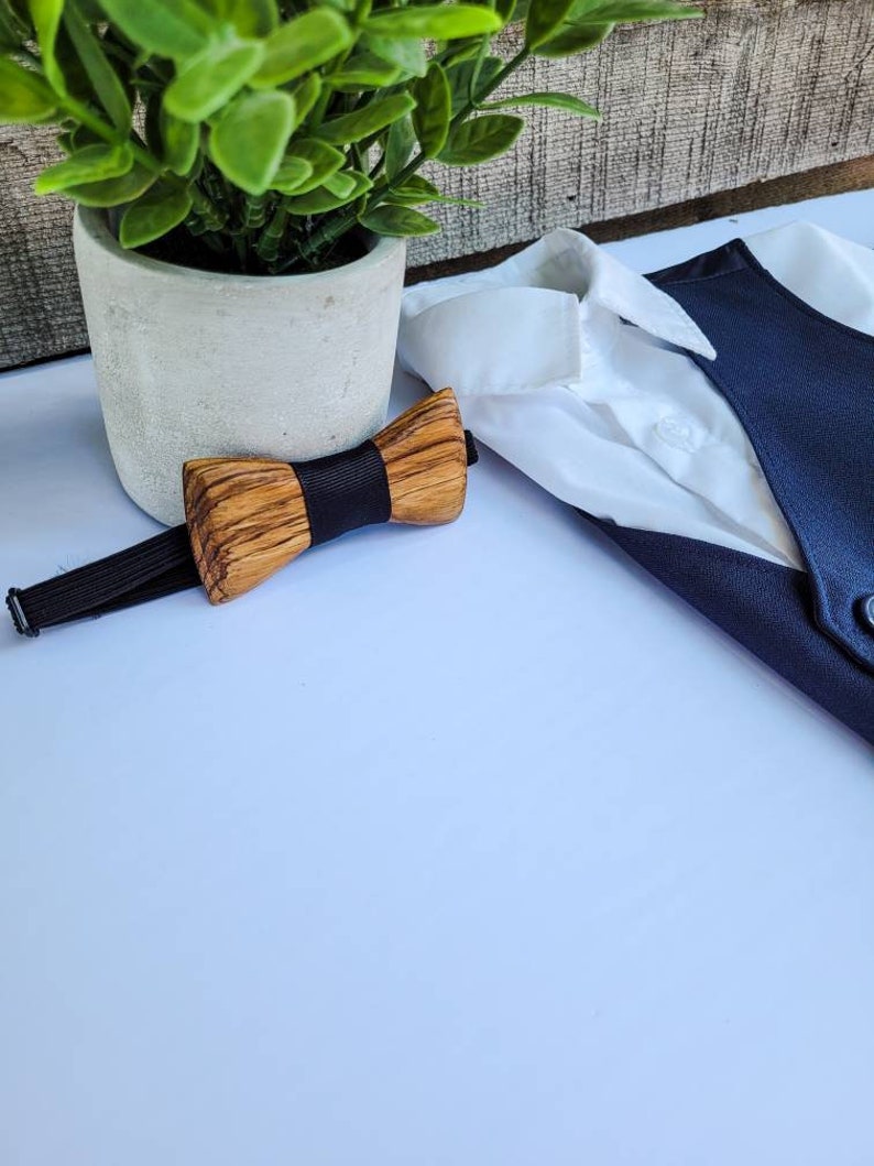 Puede incluir: Una pajarita de madera con una banda el&aacute;stica negra. La pajarita est&aacute; hecha de madera marr&oacute;n claro y tiene una veta de madera natural. La pajarita est&aacute; sobre una superficie blanca.