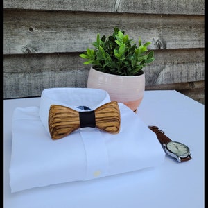 Puede incluir: Una camisa blanca con una pajarita de madera. La pajarita tiene un patr&oacute;n de veta de madera marr&oacute;n oscuro y una banda negra en el centro. La camisa est&aacute; doblada y colocada sobre una superficie blanca.