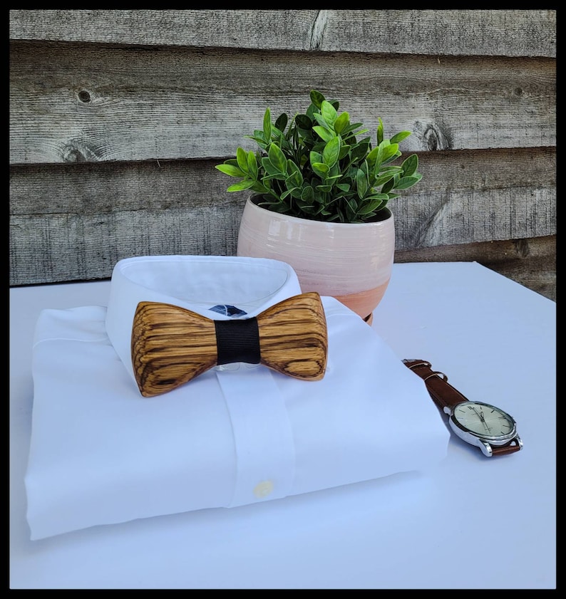 Puede incluir: Una camisa blanca con una pajarita de madera. La pajarita tiene un patr&oacute;n de veta de madera marr&oacute;n oscuro y una banda negra en el centro. La camisa est&aacute; doblada y colocada sobre una superficie blanca.