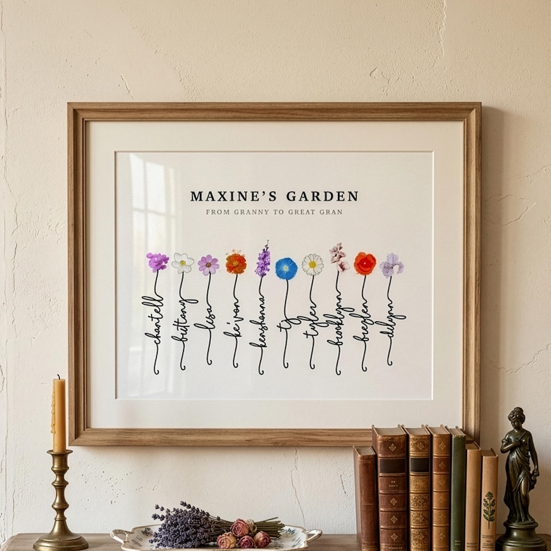 Maxine's Garden personalized birth flower print in a wood frame with white mat, hung above a table with brass candlestick, dried lavender and antique books. Subtitle reads From Granny to Great Gran. Ten watercolor birth month flowers, including violet, narcissus, cosmos, marigold, larkspur, morning glory, daisy, cherry blossom, poppy and iris, grow from hand-lettered stems spelling each name. Great grandma gift and personalized birth flower art.