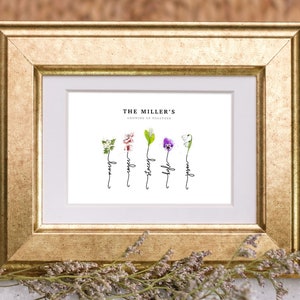 Grandmas Garden personalized birth flower print in a gold frame with white mat, sitting on a marble table with dried flowers. Five watercolor birth month flowers (hawthorn, cherry blossom, lily of the valley, violet and snowdrop) grow from hand-lettered stems that spell each name. Custom family name art and sentimental grandma gift.